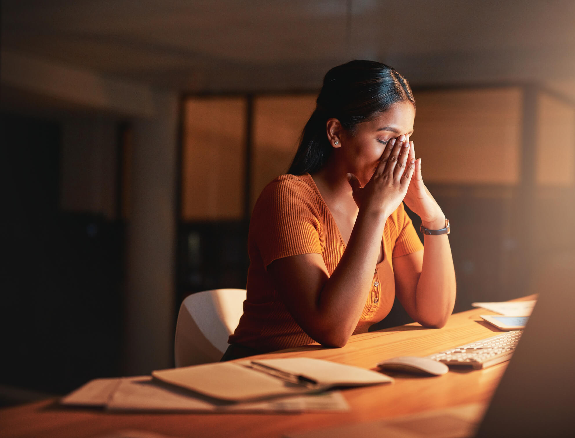 Woman in an orange shirt sitting down with her hands to her face in distress.