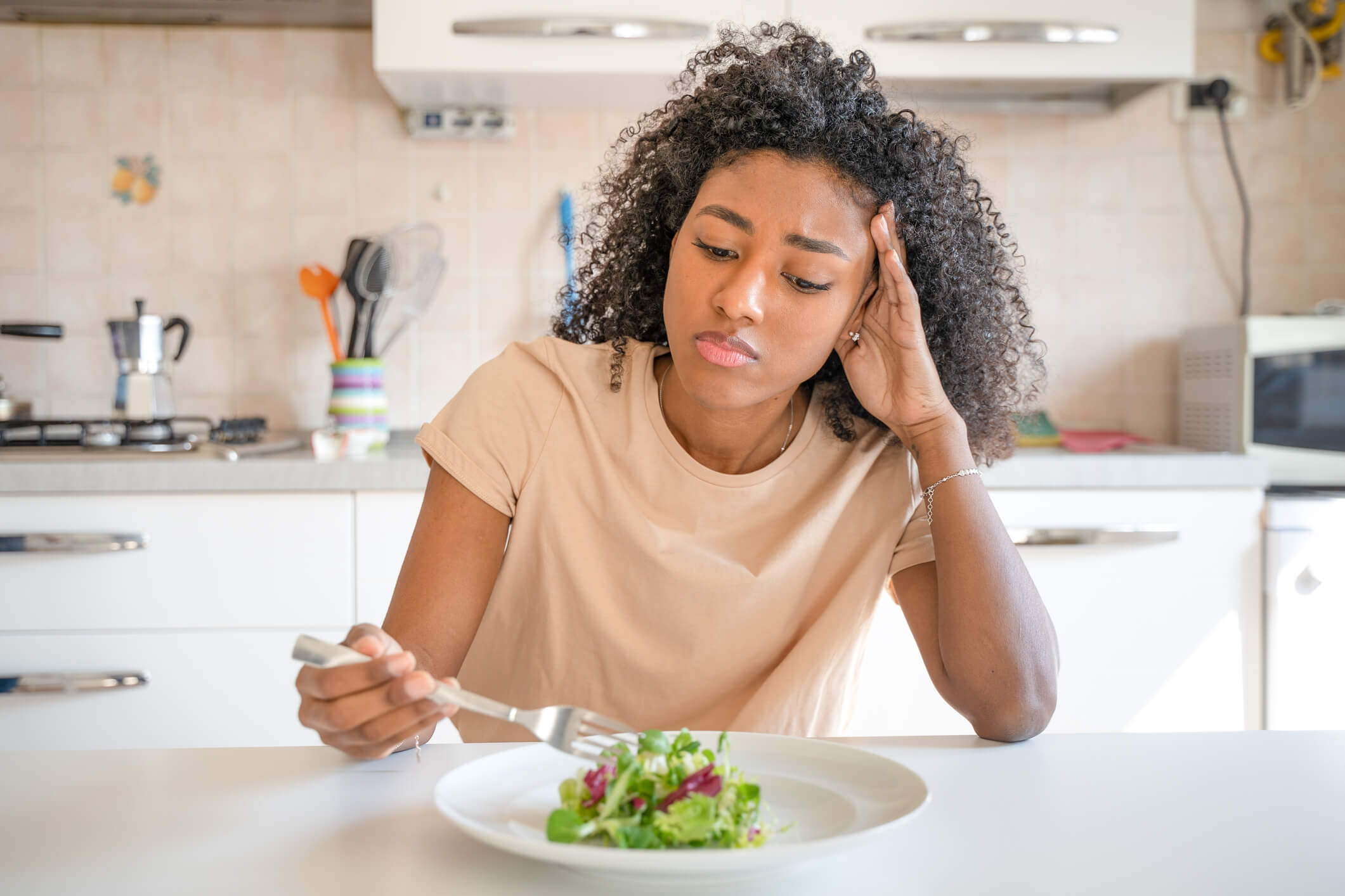 Woman with a distressed expression looking down at a salad while holding a fork and sitting at a dinner table.