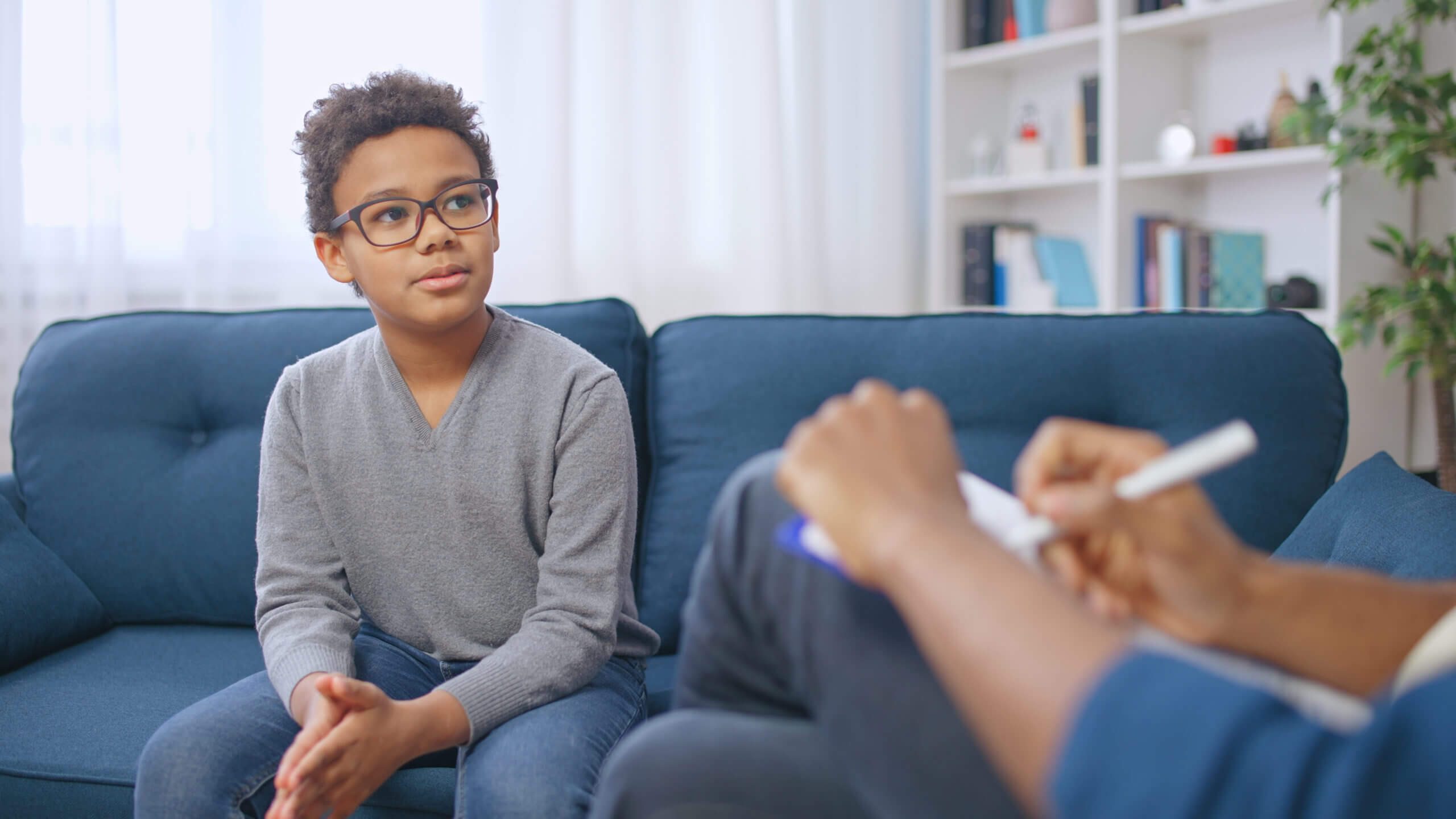 Young boy wearing glasses in a gray sweater undergoing transitions counseling with a doctor, talking about his life transitions.