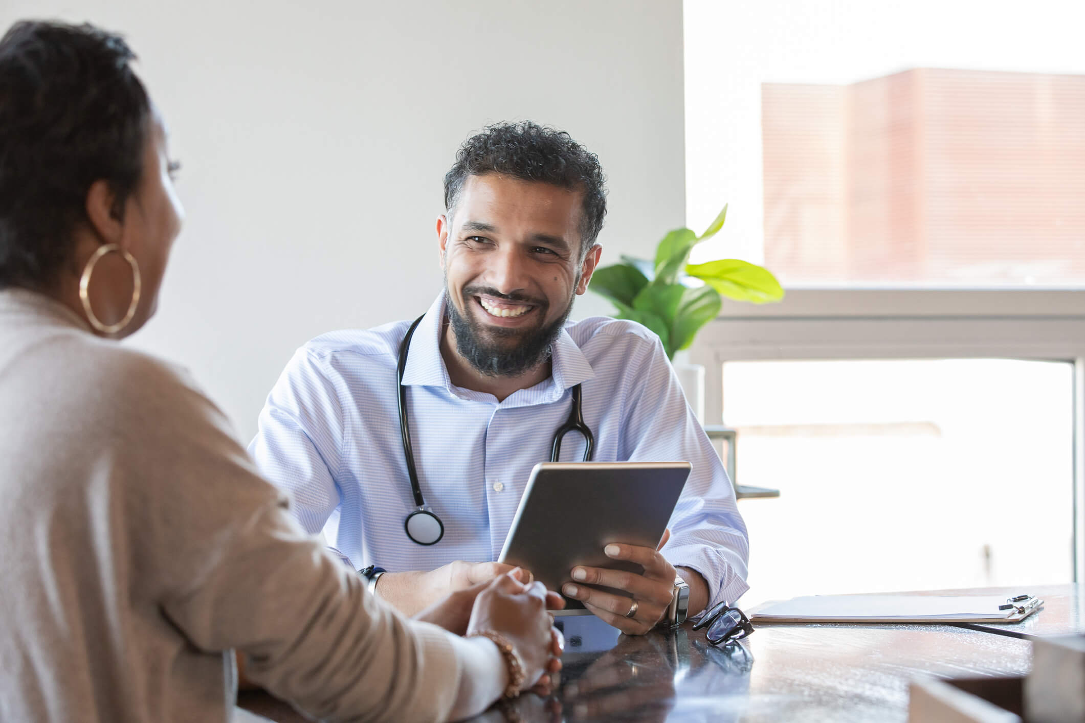 Doctor wearing a stethoscope sitting down holding a tablet smiling at a patient.