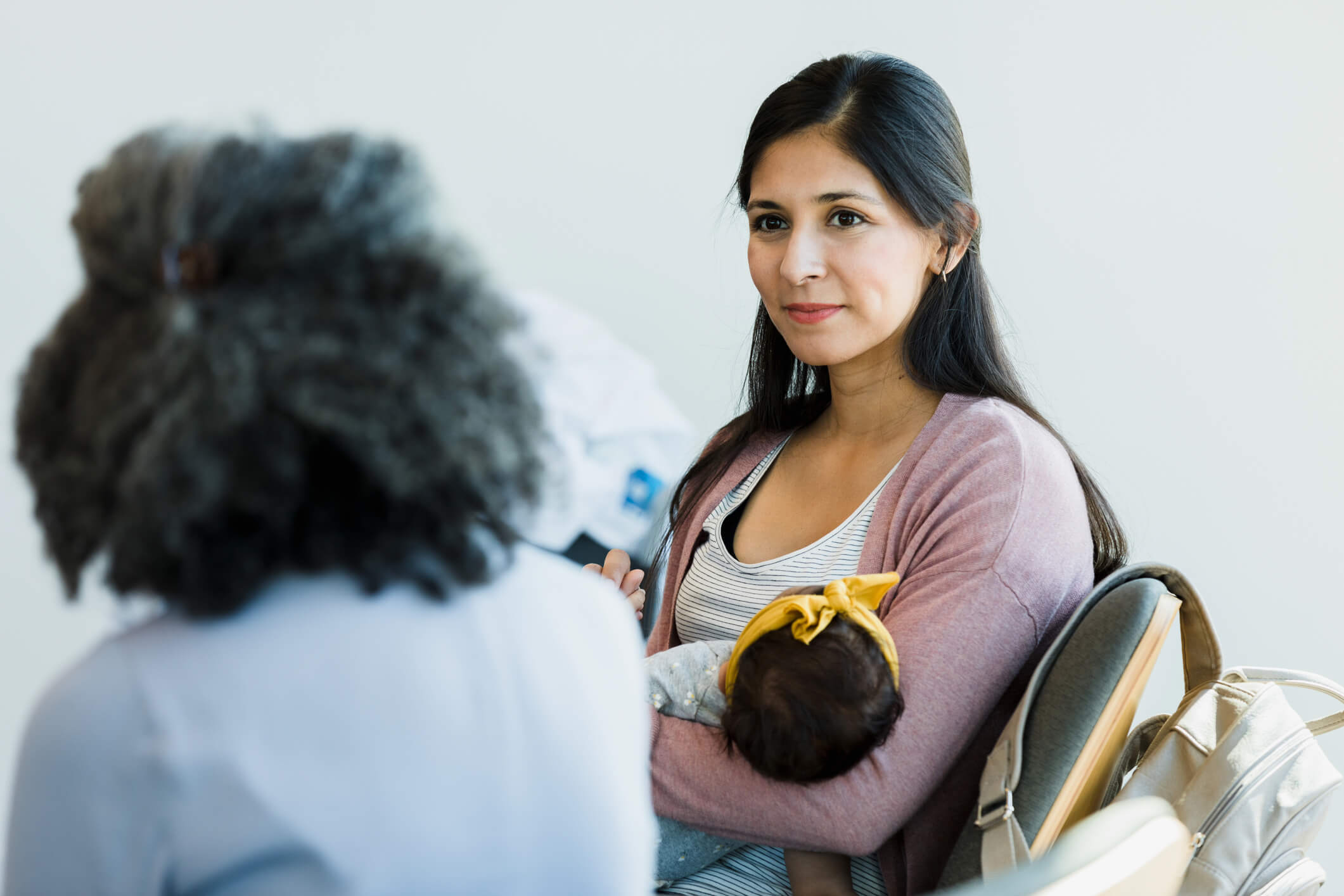 Woman holding a baby wearing a yellow bow talking to a healthcare provider.