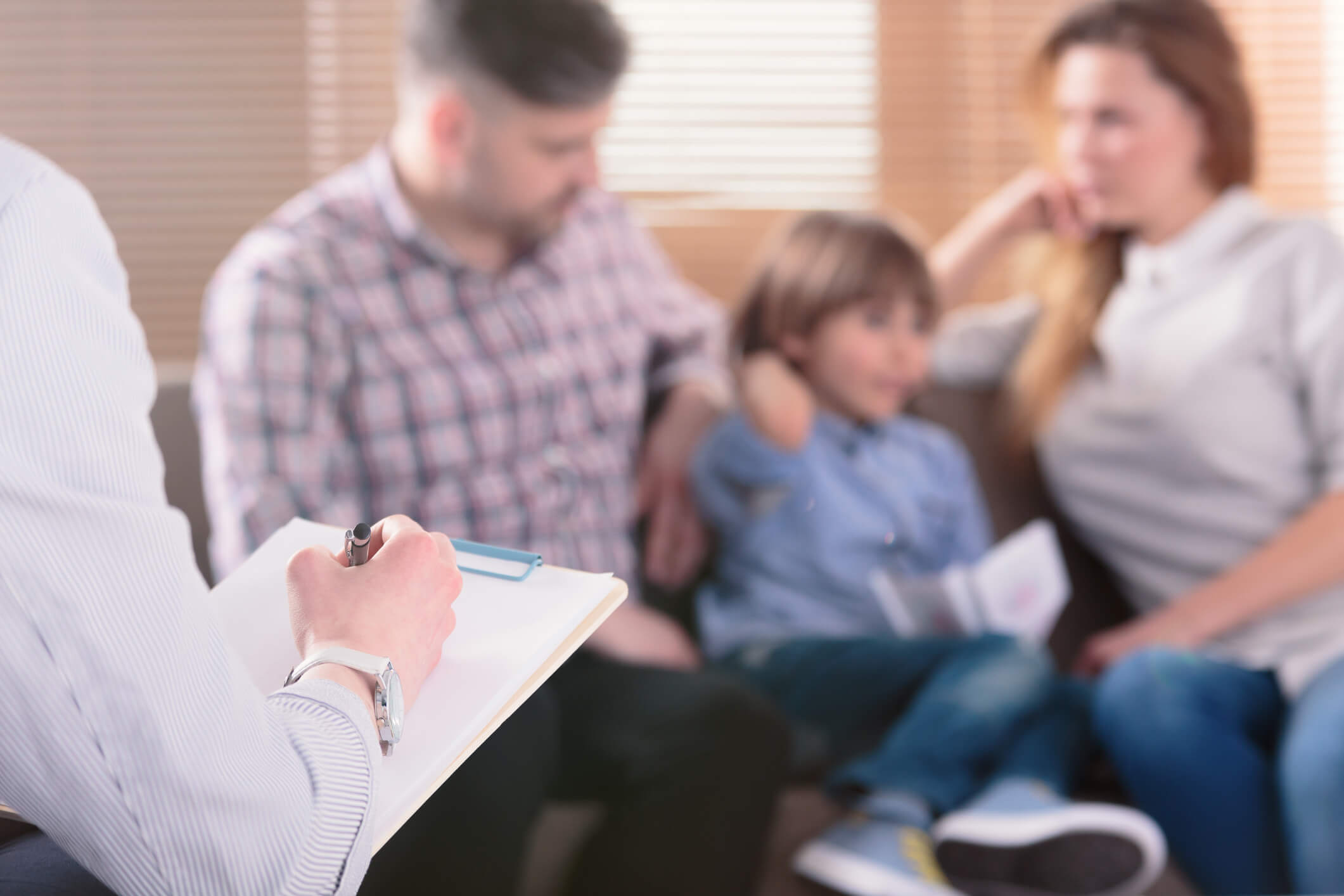 Doctor writing notes on a clipboard with a child and his family sitting on the couch in the background.