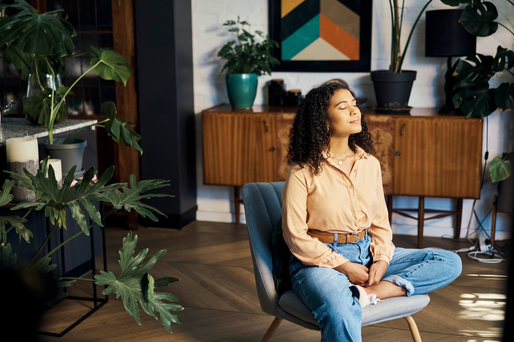 Woman in an orange button down shirt and jeans sitting in a blue chair soaking in the sunshine on her face.